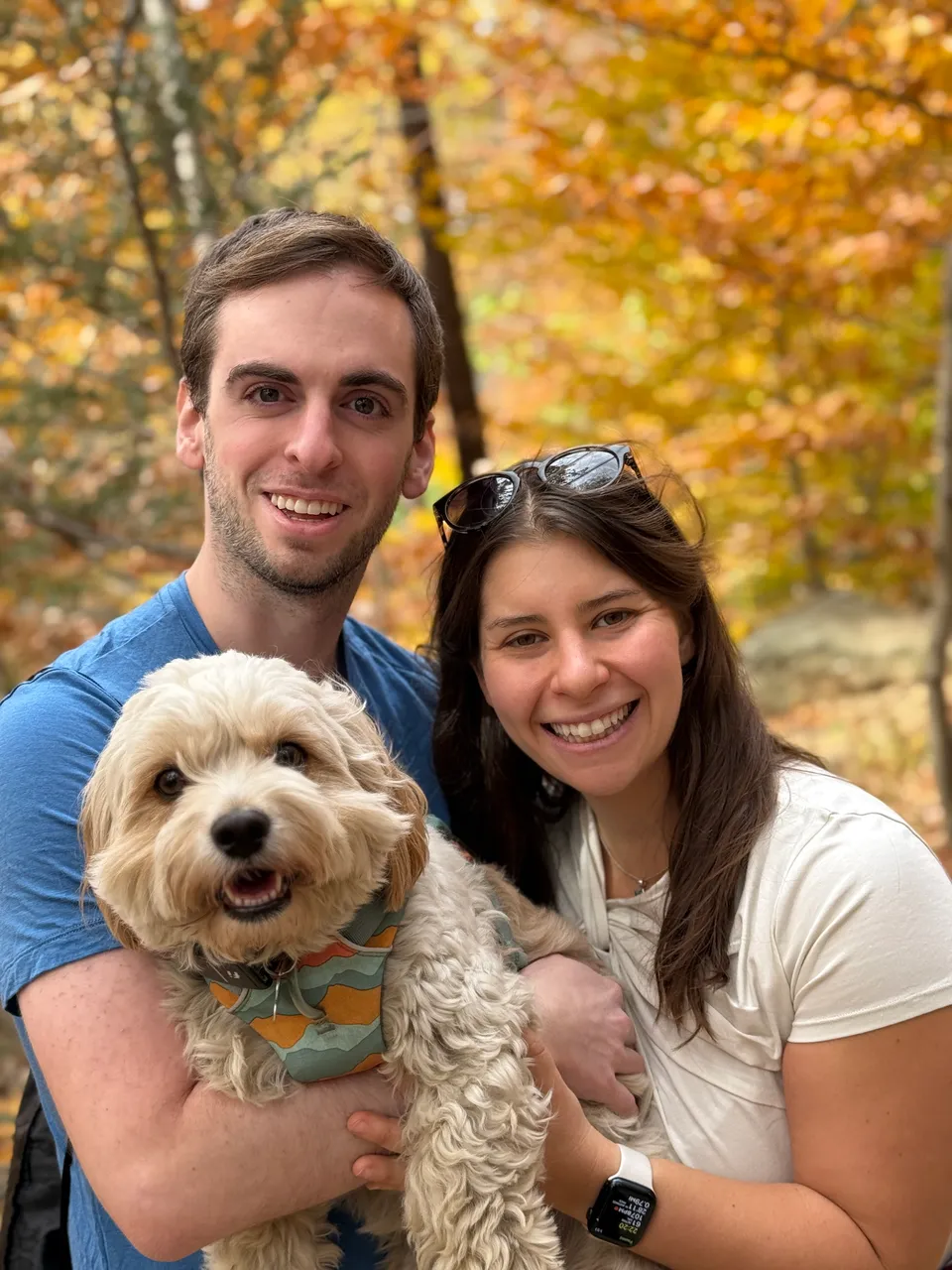 Leah and Justin holding Loki close on a fall hike, bright orange foliage behind them.