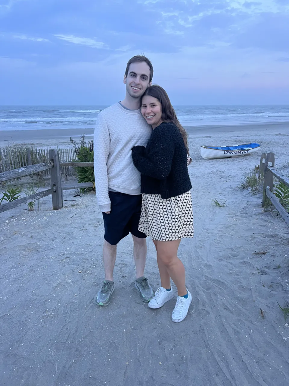 Leah and Justin hugging on the beach at dusk, dunes and a pastel evening sky behind them.