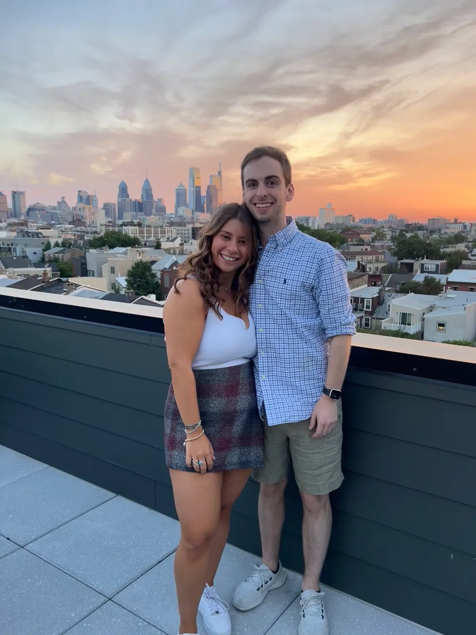 Leah and Justin arm-in-arm on a rooftop at sunset, the Philadelphia skyline glowing orange behind them.