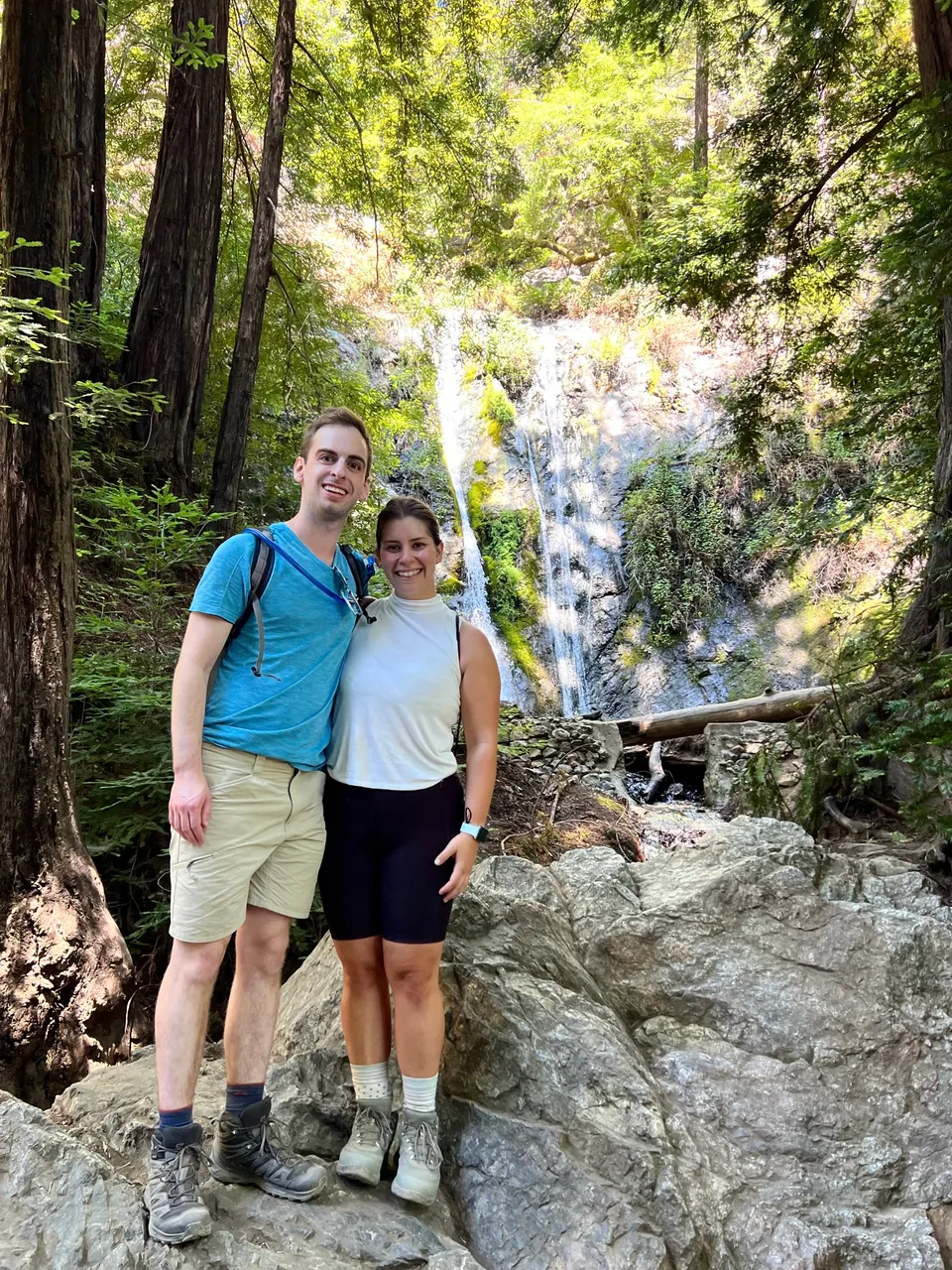 Leah and Justin in hiking gear on boulders in front of a waterfall in a redwood forest.