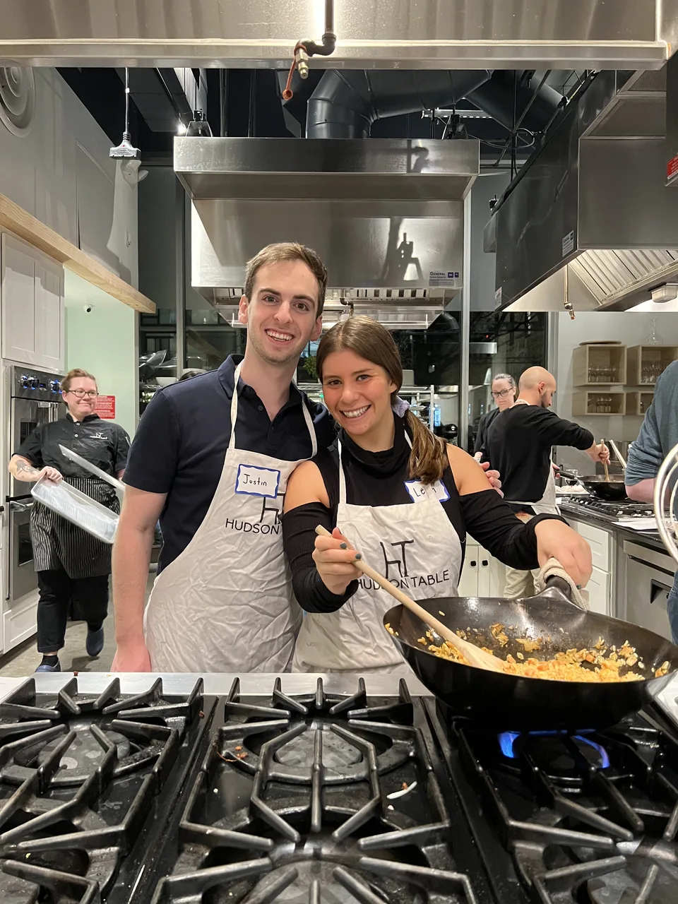 Leah and Justin in aprons at a cooking class, holding a pan of food.
