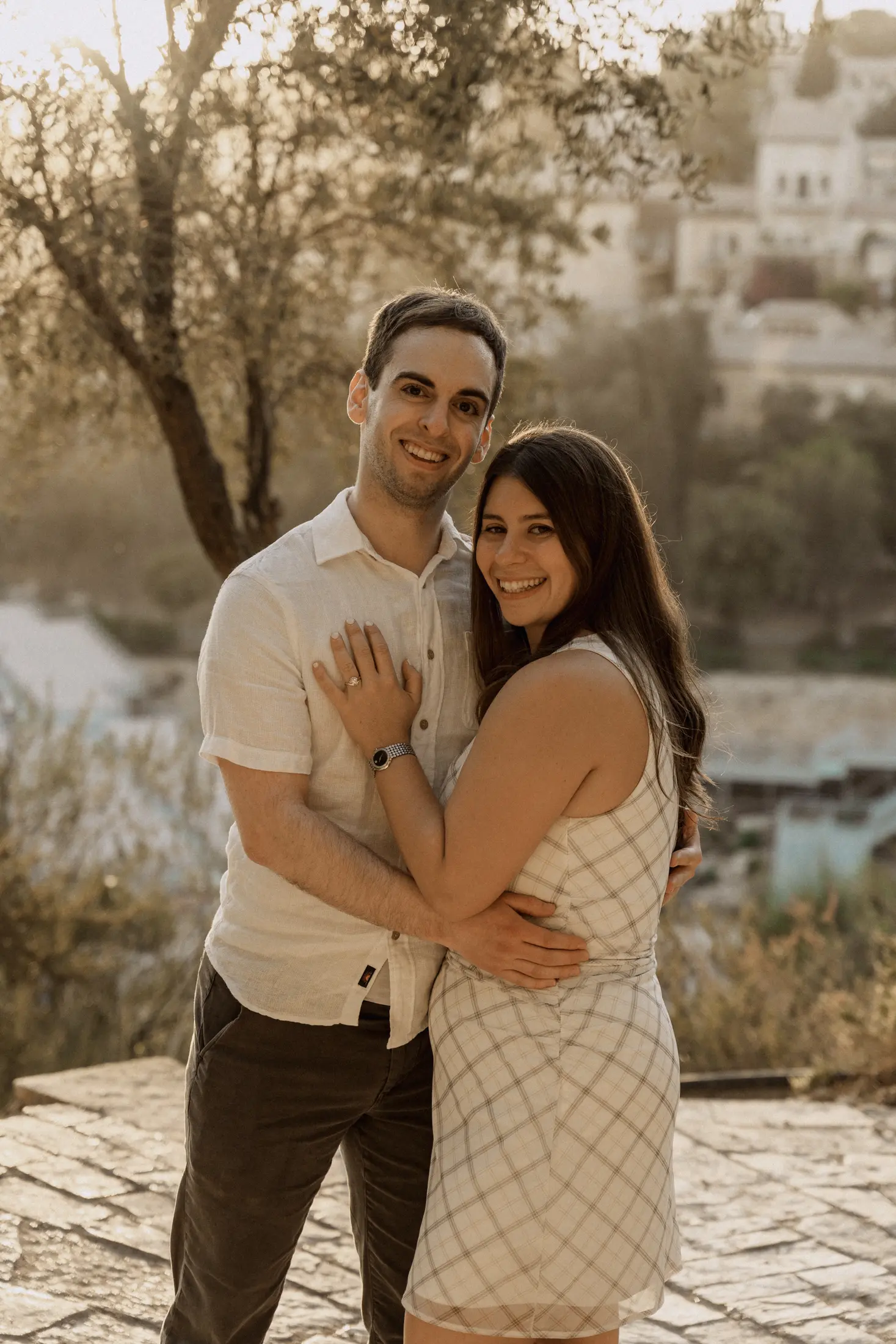 Leah and Justin embracing on a stone overlook in Jerusalem at golden hour, an olive tree and sunlit hillside behind them, Leah's ring hand resting on Justin's chest.
