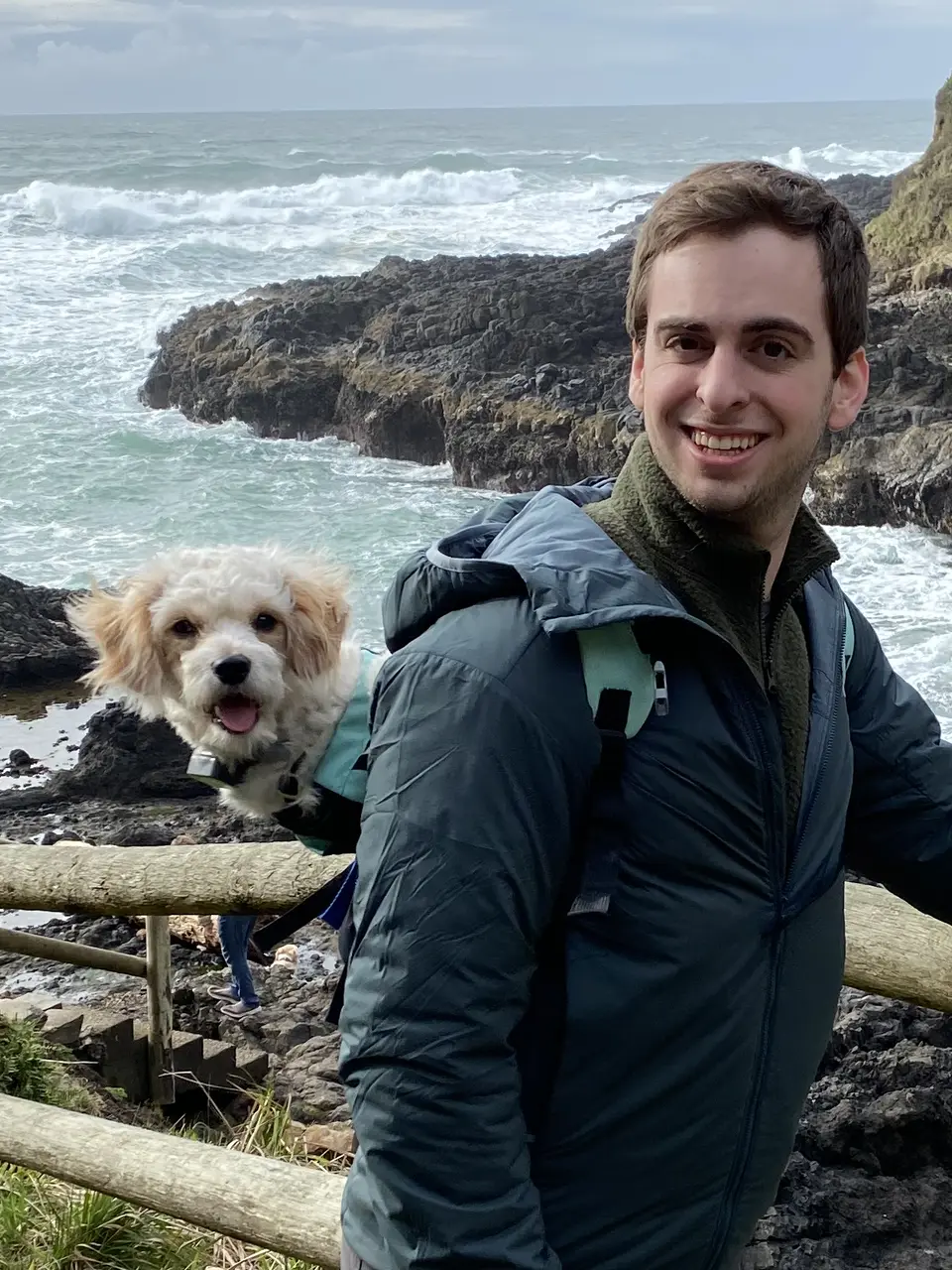 Justin sitting on a coastal fence with the sea behind him and Loki the dog nearby.