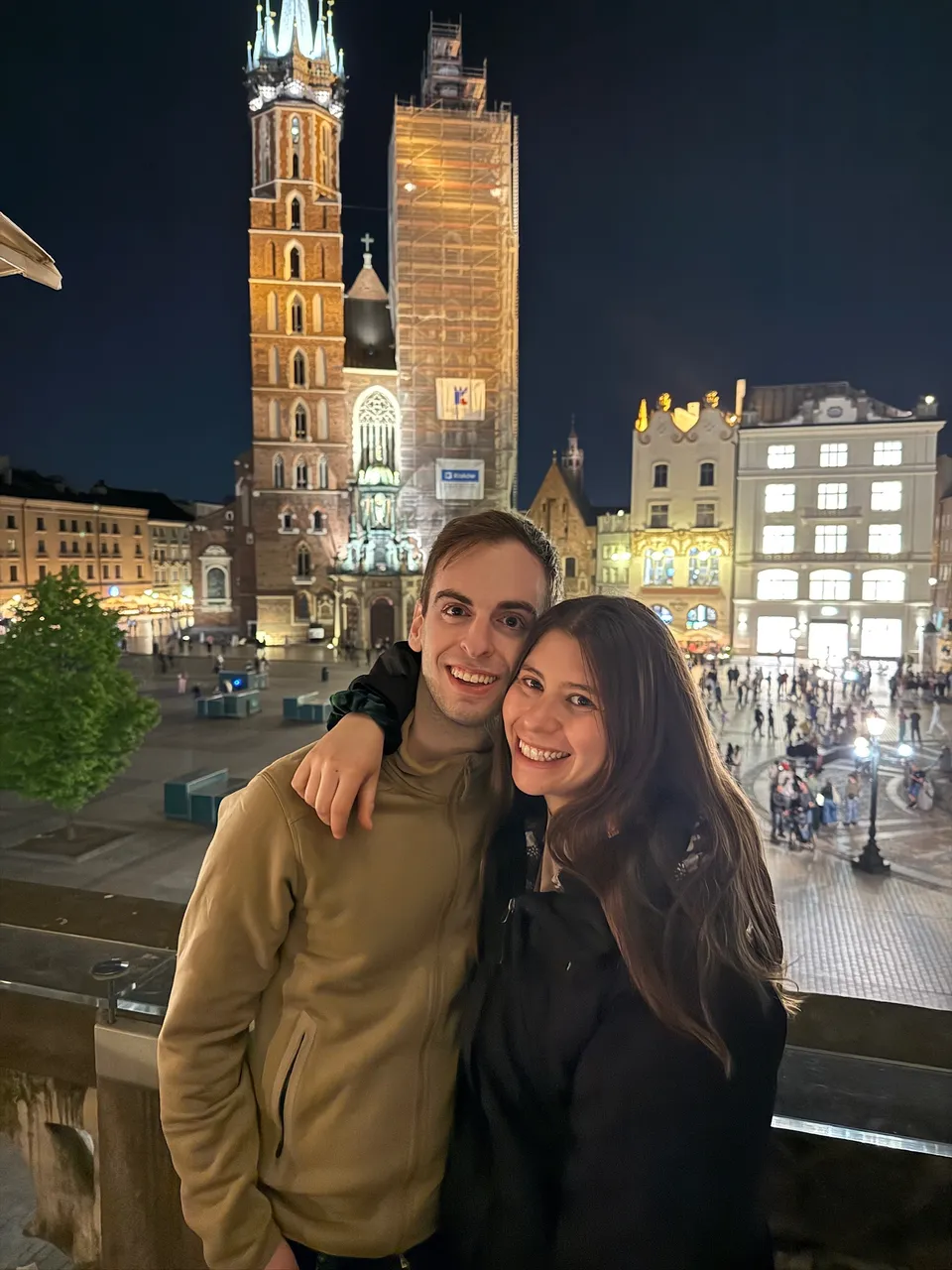 Leah and Justin on a terrace above Kraków's main square at night, St. Mary's Basilica lit gold behind them.
