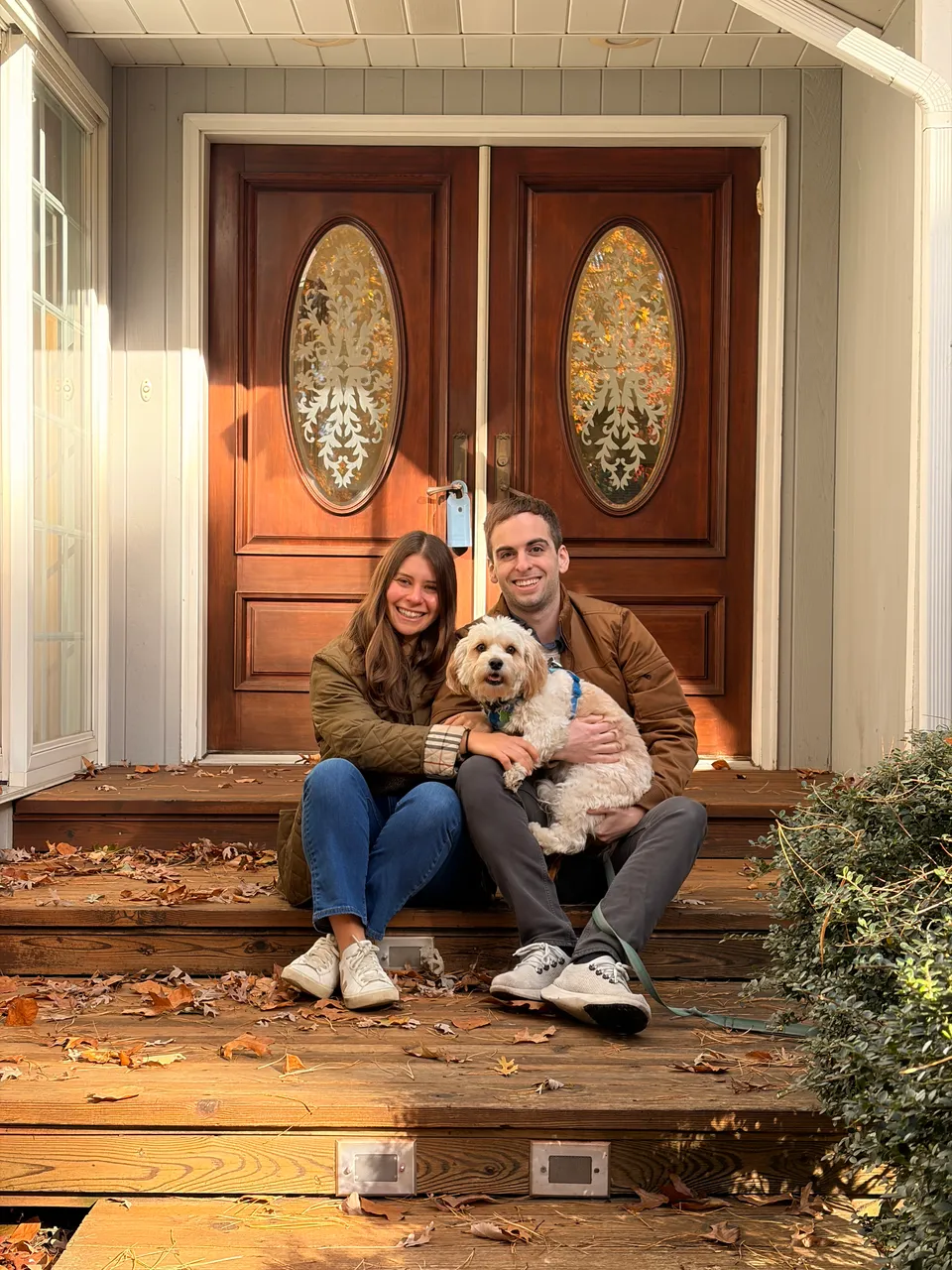 Leah and Justin on the leaf-strewn front steps of their new house, holding Loki in warm autumn light.